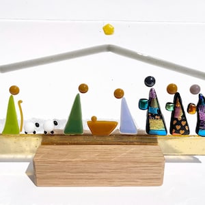 May include: A fused glass nativity scene featuring stylized figures in various colors, including green, purple, and black, arranged on a clear glass panel with a yellow star above. The piece is supported by a rectangular light wood base.