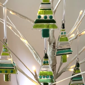 May include: Glass Christmas tree decorations in shades of green, white, and clear. The ornaments are triangular, with a square base and are suspended from white ribbons. The trees are decorated with various patterns and textures, and are hanging on a white tree.