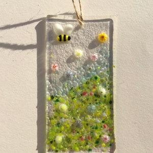 May include: A glass suncatcher featuring a bee and flowers. The bee is yellow with black stripes and is flying over a field of green, yellow, pink and white flowers.