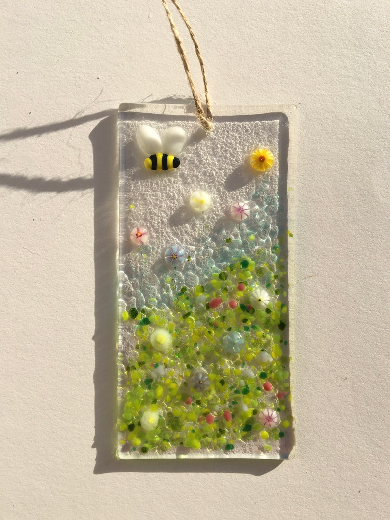May include: A glass suncatcher featuring a bee and flowers. The bee is yellow with black stripes and is flying over a field of green, yellow, pink and white flowers.