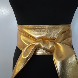 May include: A wide, gold-colored belt with a metallic sheen is tied in a bow. The belt is wrapped around a black form, showcasing its shape and design. The shiny material reflects light, highlighting the texture and color.
