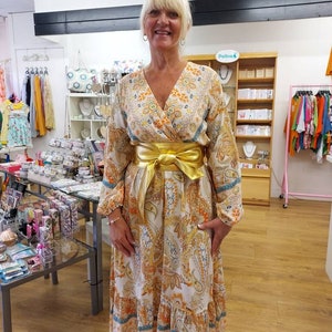May include: A long, flowing dress with a paisley pattern in shades of white, orange, and blue. The dress features long sleeves, a V-neck, and a gold-colored belt cinching the waist. The dress has a tiered skirt and is worn by a person.