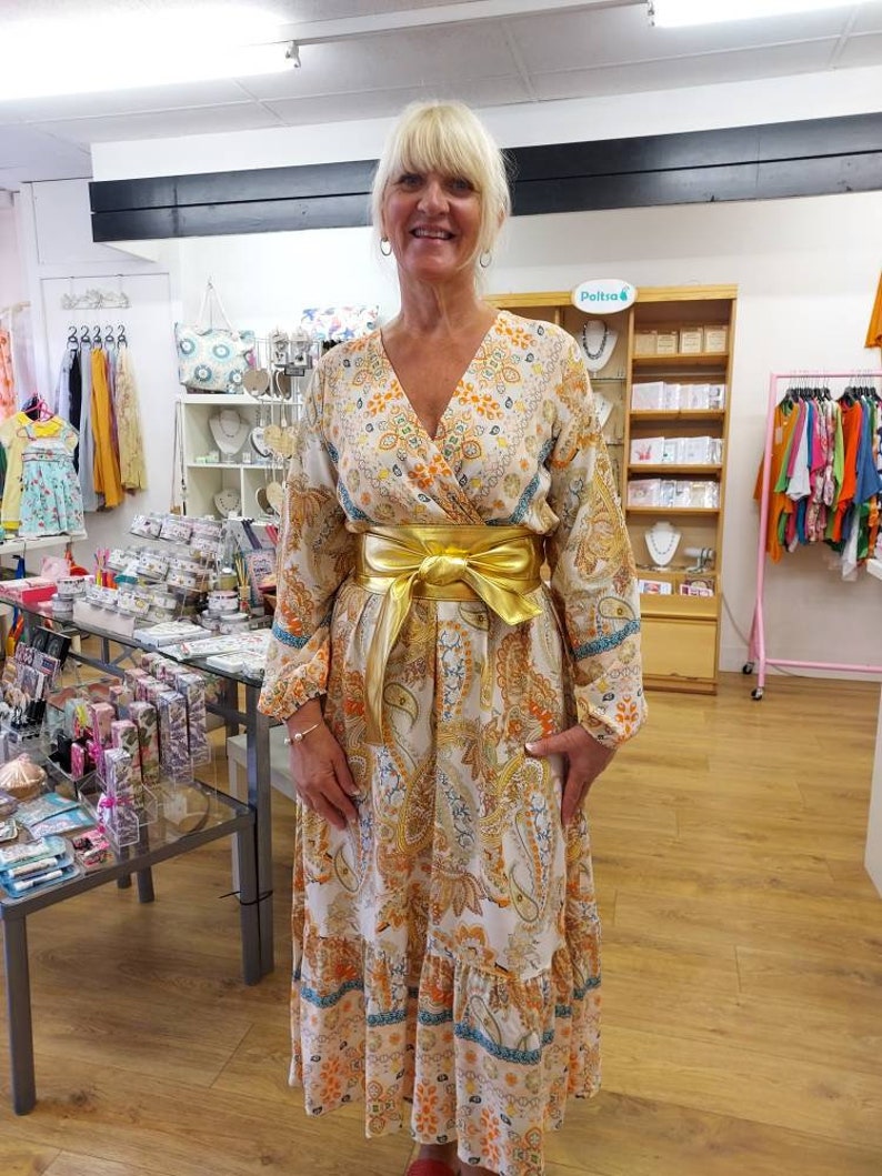 May include: A long, flowing dress with a paisley pattern in shades of white, orange, and blue. The dress features long sleeves, a V-neck, and a gold-colored belt cinching the waist. The dress has a tiered skirt and is worn by a person.