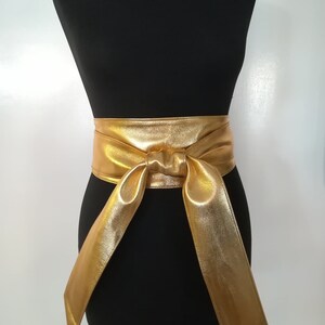 May include: A gold-colored, wide sash belt with a bow detail, displayed on a black mannequin. The belt is made of a shiny, metallic material. The sash hangs down on both sides.