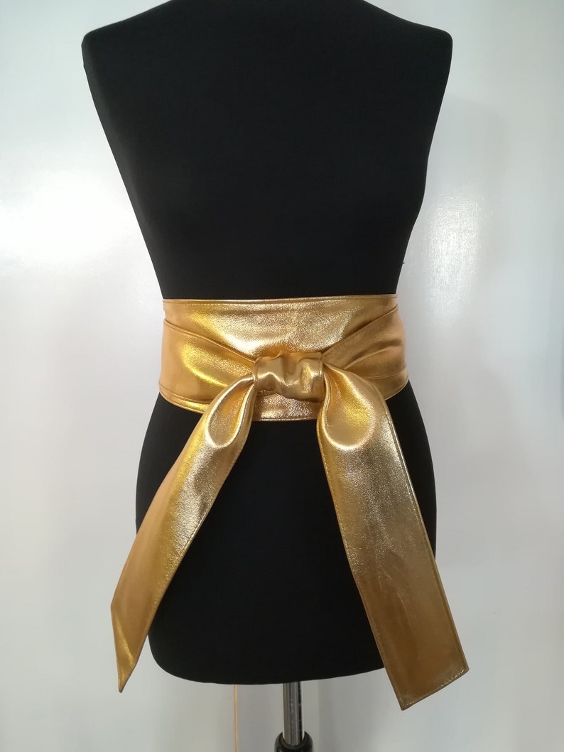 May include: A gold-colored, wide sash belt with a bow detail, displayed on a black mannequin. The belt is made of a shiny, metallic material. The sash hangs down on both sides.