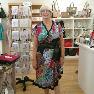 May include: A woman wearing a colorful paisley print dress with a black belt. The dress features a V-neck and short sleeves. The store background includes handbags, jewelry, and greeting cards.