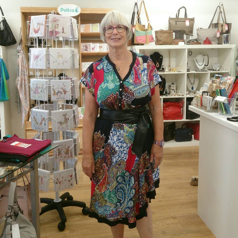 May include: A woman wearing a colorful paisley print dress with a black belt. The dress features a V-neck and short sleeves. The store background includes handbags, jewelry, and greeting cards.