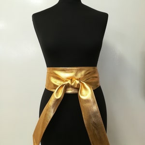 May include: A gold-colored, wide waist belt with a tied knot in the front. The belt is made of a shiny, metallic material and is displayed on a black mannequin. The belt's long ends hang down, creating a flowing effect.