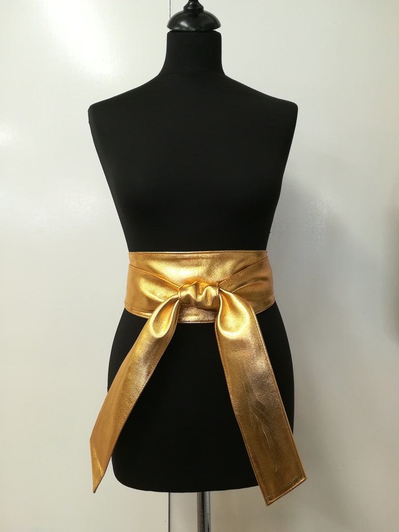 May include: A gold-colored, wide waist belt with a tied knot in the front. The belt is made of a shiny, metallic material and is displayed on a black mannequin. The belt's long ends hang down, creating a flowing effect.