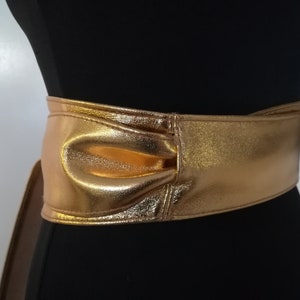 May include: A wide, gold-colored belt with a metallic sheen. The belt is wrapped around a black mannequin, showcasing its design. The belt features a bow-like detail at the front, adding a decorative element to the accessory.