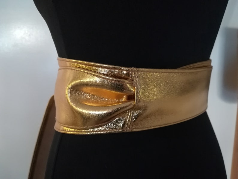 May include: A wide, gold-colored belt with a metallic sheen. The belt is wrapped around a black mannequin, showcasing its design. The belt features a bow-like detail at the front, adding a decorative element to the accessory.