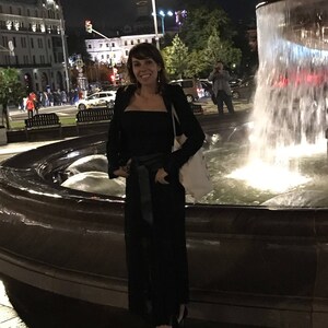 May include: A woman in a black outfit stands in front of a fountain at night. She wears a black top, pants, and jacket, with a black belt. A white tote bag hangs over her shoulder. The background includes city buildings and streetlights.