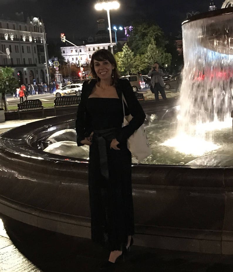 May include: A woman in a black outfit stands in front of a fountain at night. She wears a black top, pants, and jacket, with a black belt. A white tote bag hangs over her shoulder. The background includes city buildings and streetlights.