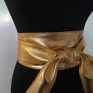 May include: A gold metallic obi belt is wrapped around a black mannequin. The belt is tied in a knot at the front, with long, flowing ends. The belt's shiny surface reflects light, creating a glamorous look.