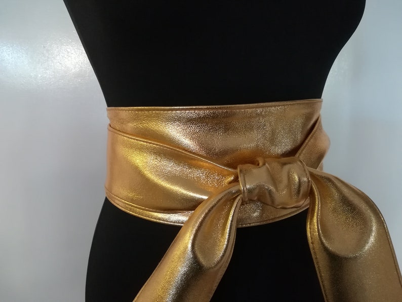 May include: A gold metallic obi belt is wrapped around a black mannequin. The belt is tied in a knot at the front, with long, flowing ends. The belt's shiny surface reflects light, creating a glamorous look.