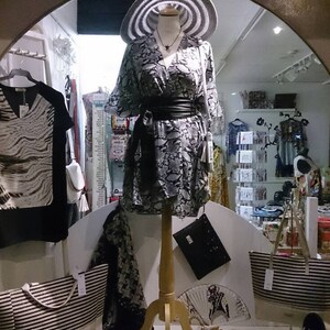 May include: A boutique window display featuring a mannequin in a patterned dress, wide-brimmed hat, and black belt. Other items include striped tote bags, a clutch, and a black and white patterned top. Accessories like jewelry and sandals are also visible.
