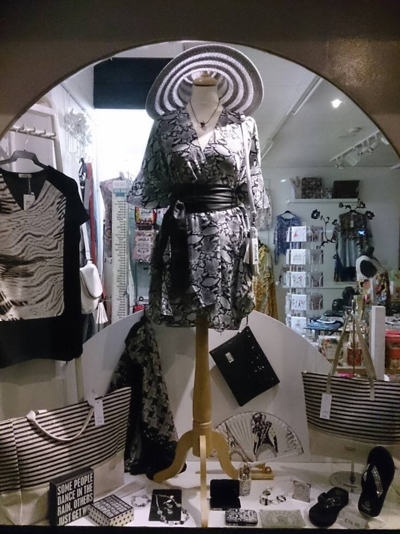 May include: A boutique window display featuring a mannequin in a patterned dress, wide-brimmed hat, and black belt. Other items include striped tote bags, a clutch, and a black and white patterned top. Accessories like jewelry and sandals are also visible.