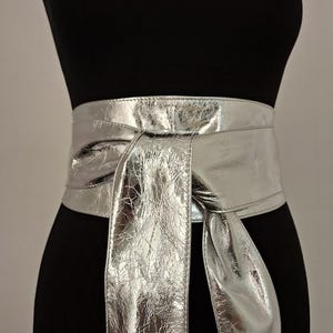 May include: A wide, metallic silver obi-style belt with a high-shine, crinkled texture. The belt features a wrap-around design that secures at the front with a long, tied knot, creating two cascading fabric ends that hang down from the centre waist.