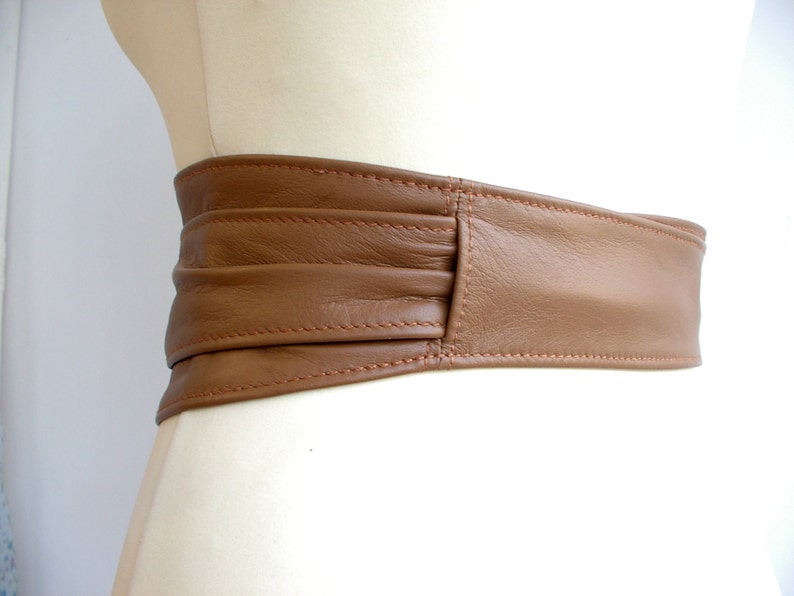 May include: A brown leather belt with a wide, folded design. The belt is stitched with a light brown thread.