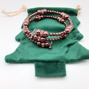 May include: A multi-strand wrap bracelet featuring dark brown spherical beads and silver-toned spacers embedded with small pink crystals. The bracelet is coiled in a circular shape and rests on a green velvet pouch shaped like a Christmas tree.