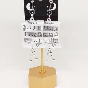 May include: A pair of silver earrings with a white background featuring musical notes and the word "Music". The earrings are hanging from a gold stand.
