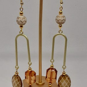 May include: A pair of gold tone dangle earrings with brown and white geometric patterned beads. The earrings feature a gold tone arch and a brown glass bead.