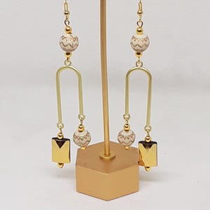 May include: Gold dangle earrings with a white and gold patterned bead and a gold square charm.