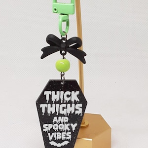 May include: Black and green glitter coffin-shaped keychain with a black bow and a green bead. The keychain has the text "Thick Thighs and Spooky Vibes" with a bat graphic.