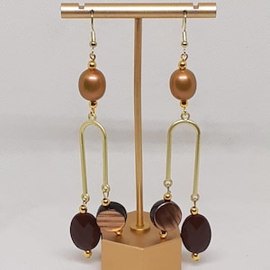 May include: Gold tone earrings with brown and wood-toned beads. The earrings feature a long, thin, gold-toned arch.