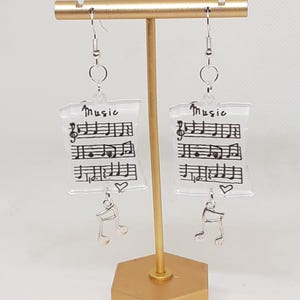 May include: A pair of silver earrings with clear square charms featuring music notes and the word "Music" written on them. Each charm has a silver music note dangling from the bottom.