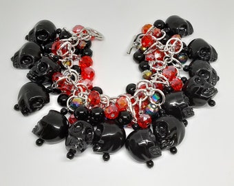 May include: A black skull charm bracelet with red and black beads. The bracelet features multiple glossy black skull charms, interspersed with red faceted beads, black round beads, and iridescent beads, all connected by a silver chain.