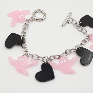 May include: A silver chain bracelet with pink glitter ghost charms and black heart charms. The bracelet has a toggle clasp. The charms are attached to the chain with small silver jump rings.
