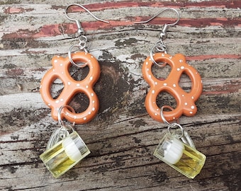 Puede incluir: Un par de pendientes con un pretzel y un abalorio de jarra de cerveza. El pretzel es naranja con puntos blancos y la jarra de cerveza es amarilla con una espuma blanca.