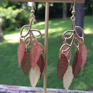K&ouml;nnte beinhalten: Ein Paar goldfarbene Ohrringe mit einem Blattdesign. Die Bl&auml;tter sind aus durchsichtigem Kunststoff in Braun-, Rosa- und Cremet&ouml;nen.