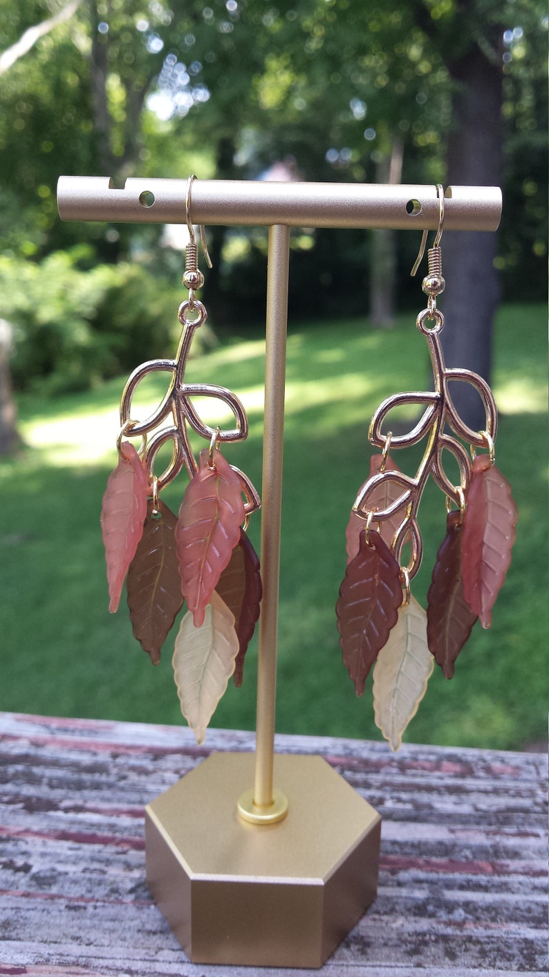 K&ouml;nnte beinhalten: Ein Paar goldfarbene Ohrringe mit einem Blattdesign. Die Bl&auml;tter sind aus durchsichtigem Kunststoff in Braun-, Rosa- und Cremet&ouml;nen.