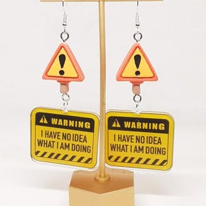 May include: A pair of novelty earrings featuring a yellow and orange warning sign design. Each earring has a triangular warning sign with an exclamation point above a rectangular sign with the text "WARNING I HAVE NO IDEA WHAT I AM DOING."