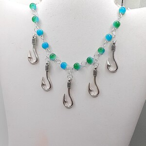 May include: A silver-toned necklace featuring a chain of blue and green beads. Dangling from the chain are five silver-colored fish hooks. The necklace is displayed on a white mannequin bust.
