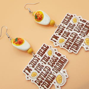 May include: A pair of novelty earrings featuring a deviled egg design with a white base, orange yolk, and green garnish. Each earring has a charm with the text "I'M JUST HERE FOR THE DEVILED EGGS & FAMILY DRAMA."