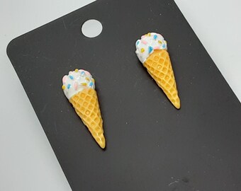 May include: Two ice cream cone earrings. The cones are a light yellow with a waffle pattern. The ice cream is white with pink, blue, and yellow sprinkles. The earrings are on a black background.
