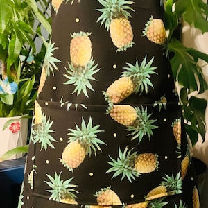 May include: Black apron with a pineapple print and white polka dots. The apron has a large pocket in the front.