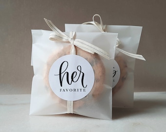 Peut inclure: Deux sacs en papier blanc avec un ruban blanc noué autour. Le sac de gauche a un autocollant rond avec le texte "her favorite" en écriture noire.