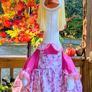 May include: A decorative goose figurine with a white body, orange beak, and yellow yarn hair. It wears a pink dress with ribbon patterns and a matching bow. The goose is standing on a wooden surface with fall foliage in the background.