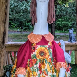 May include: A whimsical goose statue wearing a sunflower print dress, orange polka dot collar, and a matching bow. The goose has brown yarn hair and a painted orange beak. The statue is a decorative garden ornament.