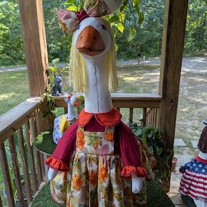 May include: A whimsical goose figurine wearing a fall-themed dress with orange and yellow leaf patterns. The goose has a white head, an orange beak, and blonde hair. A matching bow adorns the head. The figurine is on a green surface.