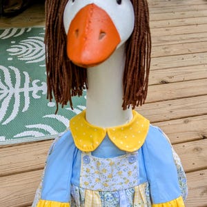 May include: A white goose doll wearing a blue dress with yellow accents and a yellow and blue bow. The dress has a floral pattern and a white lace trim. The goose has brown yarn hair.