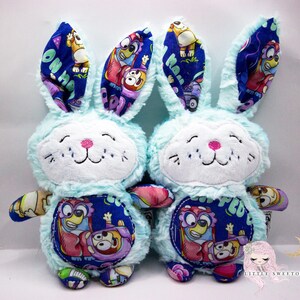 May include: Two blue plush bunny toys with embroidered faces. The bunnies have colorful fabric ears with cartoon characters on them. The fabric on the bunnies' bodies is blue with a cartoon character on the front.