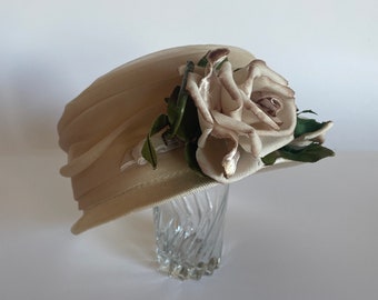 May include: A beige hat with a large white rose on top. The hat is made of a soft, flowing fabric and has a wide brim.