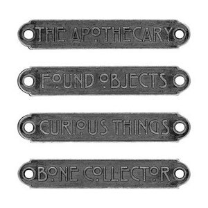 May include: Four rectangular, dark gray metal signs with rounded edges and holes for mounting. Each sign features embossed white text: "THE APOTHECARY", "FOUND OBJECTS", "CURIOUS THINGS", and "BONE COLLECTOR". The signs are arranged vertically against a white background.