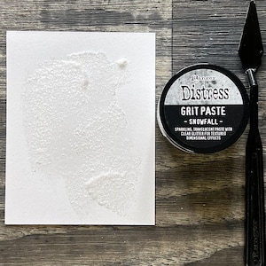 May include: A white textured paste application on a white card, next to a jar of Ranger Distress Grit Paste - Snowfall, and a black palette knife. The jar is black with white text that reads "Sparkling, translucent paste with clear glitter for textured dimensional effects."