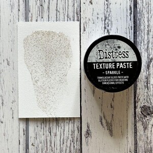 May include: A black jar of Ranger Distress Texture Paste in Sparkle. The jar has a white label with the text "Ranger Distress Texture Paste - Sparkle - Translucent gloss paste with glitter flecks for creating dimensional effects."
